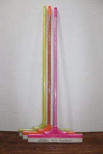 Three squeegees are lined up against a plain wall on a wooden surface. Each has a brightly colored handle in neon shades of yellow, orange, and pink. The squeegee blades and handles are wrapped in clear plastic packaging. The base of each squeegee has the branding 'Cleanux Wiper Industry' and 'King Master' printed on it.