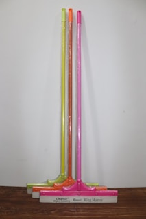 Three squeegees are lined up against a plain wall on a wooden surface. Each has a brightly colored handle in neon shades of yellow, orange, and pink. The squeegee blades and handles are wrapped in clear plastic packaging. The base of each squeegee has the branding 'Cleanux Wiper Industry' and 'King Master' printed on it.