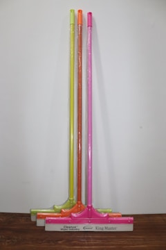 Three squeegees are lined up against a plain wall on a wooden surface. Each has a brightly colored handle in neon shades of yellow, orange, and pink. The squeegee blades and handles are wrapped in clear plastic packaging. The base of each squeegee has the branding 'Cleanux Wiper Industry' and 'King Master' printed on it.