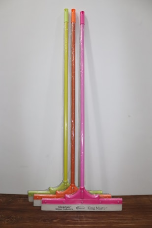 Three squeegees are lined up against a plain wall on a wooden surface. Each has a brightly colored handle in neon shades of yellow, orange, and pink. The squeegee blades and handles are wrapped in clear plastic packaging. The base of each squeegee has the branding 'Cleanux Wiper Industry' and 'King Master' printed on it.
