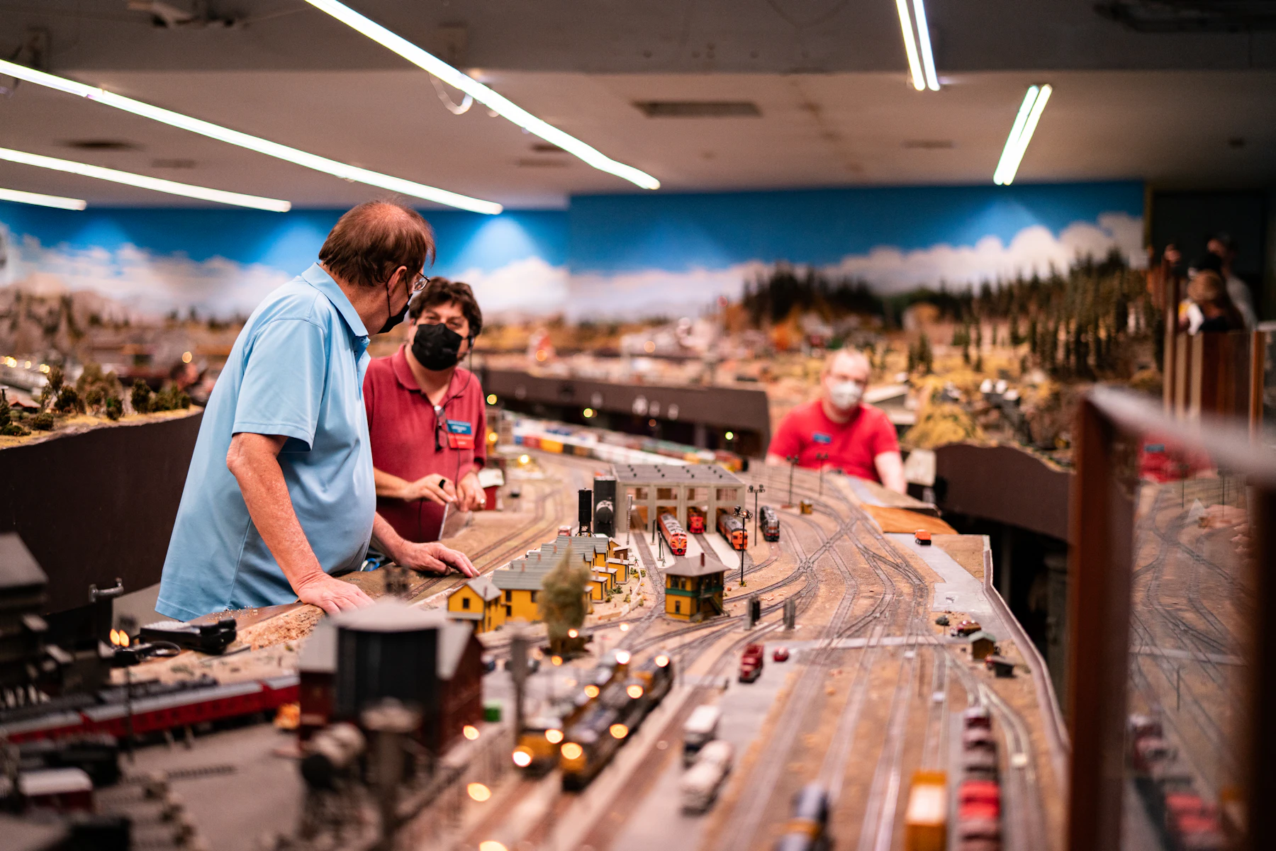 Three people standing beside a large club layout with a rail yard, buildings and a painted sky backdrop