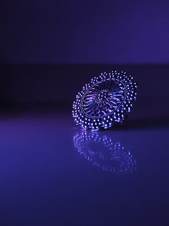A detailed, intricate piece of jewelry is positioned on a reflective surface, illuminated by purple and blue lighting. The intricate patterns and symmetry are highlighted by the lighting, creating an elegant and captivating reflection.