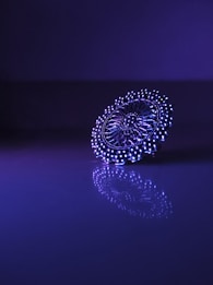 A detailed, intricate piece of jewelry is positioned on a reflective surface, illuminated by purple and blue lighting. The intricate patterns and symmetry are highlighted by the lighting, creating an elegant and captivating reflection.
