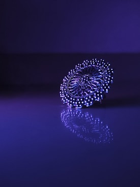 A detailed, intricate piece of jewelry is positioned on a reflective surface, illuminated by purple and blue lighting. The intricate patterns and symmetry are highlighted by the lighting, creating an elegant and captivating reflection.