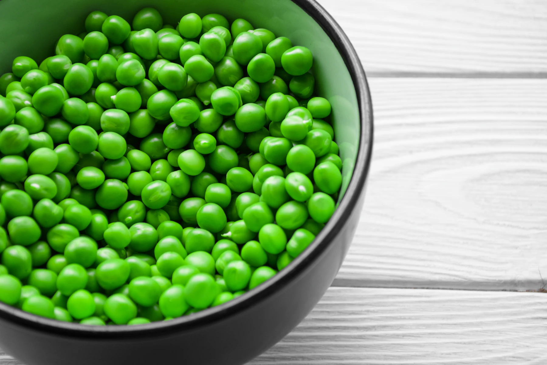 A dark bowl full of bright green peas on white wooden boards