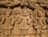 An intricate wooden carving showcasing three figures in traditional attire. The figures are adorned with detailed headgear and garments, holding various postures indicative of cultural significance. The background features elaborate carvings of patterns and what appear to be other smaller figures.