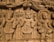 An intricate wooden carving showcasing three figures in traditional attire. The figures are adorned with detailed headgear and garments, holding various postures indicative of cultural significance. The background features elaborate carvings of patterns and what appear to be other smaller figures.