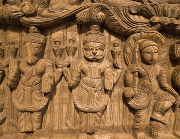 An intricate wooden carving showcasing three figures in traditional attire. The figures are adorned with detailed headgear and garments, holding various postures indicative of cultural significance. The background features elaborate carvings of patterns and what appear to be other smaller figures.