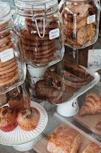 Assortment of baked goods displayed in a glass bakery case. Large glass jars contain stacks of cookies, including chocolate and oatmeal raisin varieties. Muffins with raisins are placed on a plate in the foreground, while croissants and pastries are arranged on wooden boards.