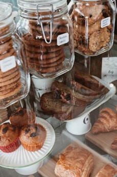 Assortment of baked goods displayed in a glass bakery case. Large glass jars contain stacks of cookies, including chocolate and oatmeal raisin varieties. Muffins with raisins are placed on a plate in the foreground, while croissants and pastries are arranged on wooden boards.