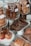 Assortment of baked goods displayed in a glass bakery case. Large glass jars contain stacks of cookies, including chocolate and oatmeal raisin varieties. Muffins with raisins are placed on a plate in the foreground, while croissants and pastries are arranged on wooden boards.