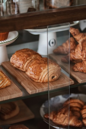 A display case featuring a variety of pastries, including croissants and chocolate-filled pastries, arranged on wooden trays. The pastries have a golden-brown, flaky appearance with a shiny glaze on top.