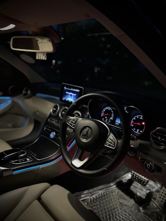 A luxurious car interior featuring a sleek black steering wheel with a silver logo, illuminated gauges on the dashboard, and an integrated infotainment screen displaying digital content. The design includes leather upholstery and ambient lighting with blue accents. The rearview mirror and other standard controls are visible, all within a well-crafted and modern aesthetic space.