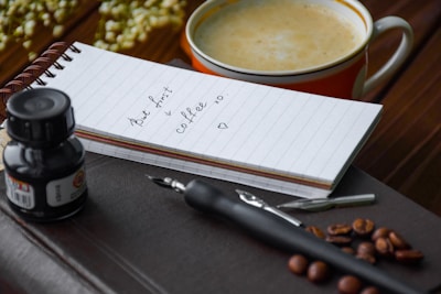 A small notebook with a handwritten message sits on top of a leather-bound book. Next to it, a black ink bottle and fountain pen are placed. On the right, a cup filled with coffee is partially visible. Coffee beans are scattered around the scene, hinting at a warm, cozy atmosphere.