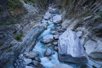 A narrow mountain gorge with a fast-flowing stream. The water appears clear and has a bluish hue, twisting around large grey and blue-grey boulders. The rocky walls of the canyon are steep and covered with sparse vegetation, giving a rugged and natural feel.
