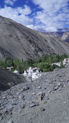 A mountainous landscape with rocky terrain in the foreground and a lush green area with trees in the middle ground. Several white stupa-like structures are visible amidst the greenery, near a small cluster of buildings. The sky is partially cloudy with blue patches, and the mountains in the background are rugged and bare.