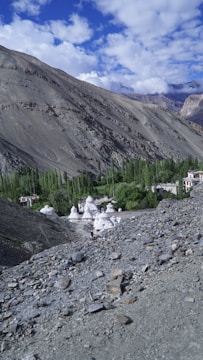 A mountainous landscape with rocky terrain in the foreground and a lush green area with trees in the middle ground. Several white stupa-like structures are visible amidst the greenery, near a small cluster of buildings. The sky is partially cloudy with blue patches, and the mountains in the background are rugged and bare.