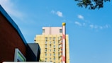 A multi-story building with a yellow facade and numerous windows, each having an air conditioning unit attached. The sky is clear with a few scattered clouds. In the foreground, there is a parking barrier with red and white stripes. Part of a brown brick wall and some tree branches are visible along the edges.