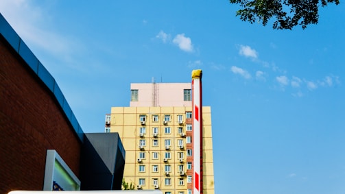 A multi-story building with a yellow facade and numerous windows, each having an air conditioning unit attached. The sky is clear with a few scattered clouds. In the foreground, there is a parking barrier with red and white stripes. Part of a brown brick wall and some tree branches are visible along the edges.
