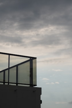 A modern glass railing on a rooftop stands silhouetted against a cloudy sky. The clouds are dense and cover much of the sky, giving it a dramatic appearance.