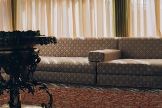 A living room setting featuring a patterned beige sofa with circular designs against a backdrop of tall, pleated curtains allowing soft natural light to filter in. In the foreground, a dark, ornately designed table or plant stand adds contrast to the lighter elements.
