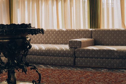 A living room setting featuring a patterned beige sofa with circular designs against a backdrop of tall, pleated curtains allowing soft natural light to filter in. In the foreground, a dark, ornately designed table or plant stand adds contrast to the lighter elements.