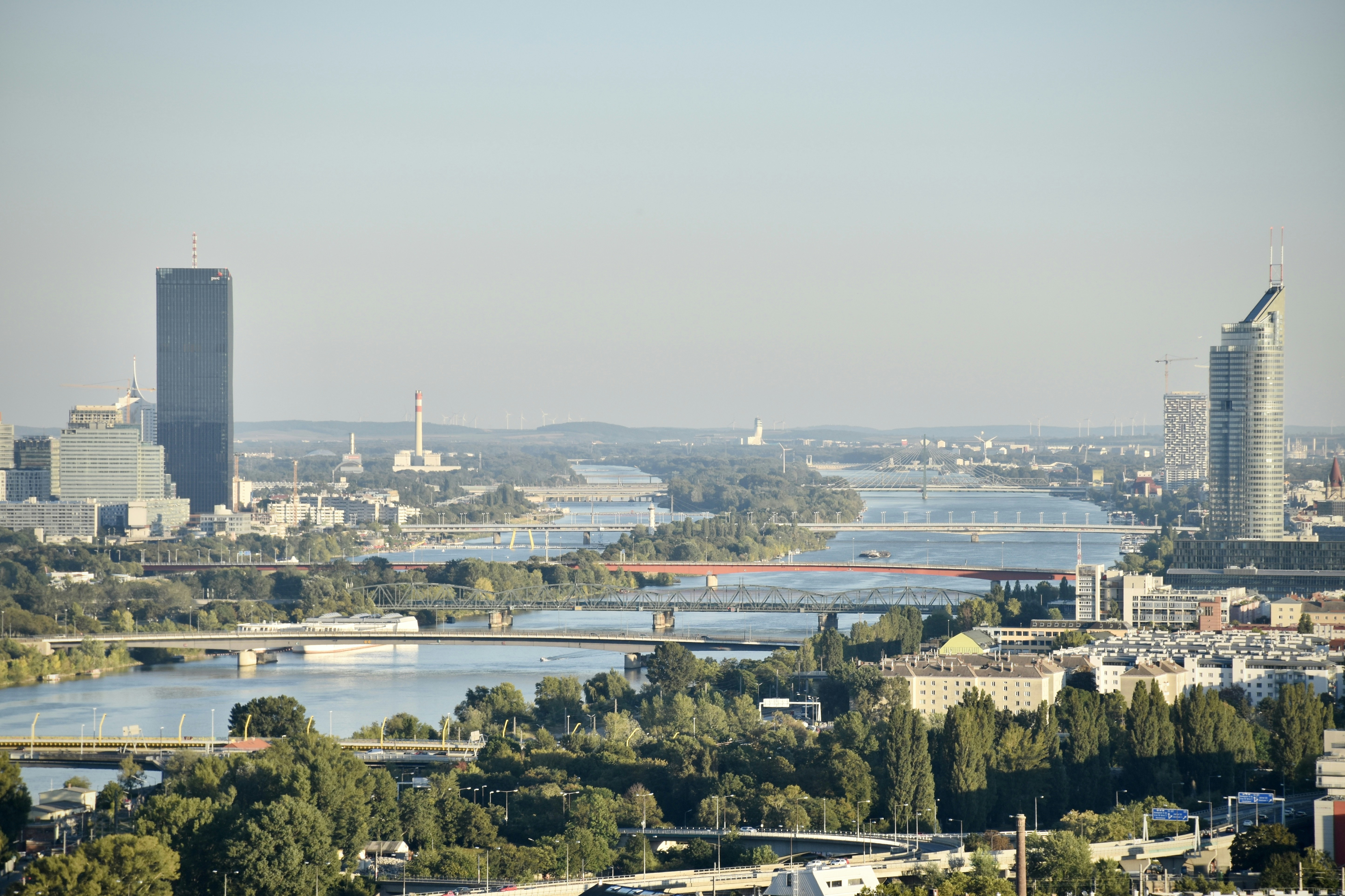 a view of a city with a river running through it, The river Danube and its bridges and high rise buildings as seen from the Döbling hills in Vienna. In the upper center of the pic you can see the Tower of Viennas Schwechat airport, distant around 20 km from where the shot was taken.