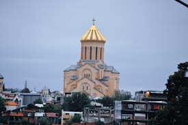A large Orthodox church with a prominent golden dome stands amidst an urban setting. The building features multiple arched windows and a detailed facade. Surrounding the church are various residential and commercial buildings. The sky is overcast, creating a serene and subdued atmosphere.