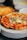 A freshly baked pizza topped with green olives, red bell pepper strips, pieces of sausage or meat, and an egg at the center. The pizza has a golden-brown crust and is placed on a stack of white plates. A blurred background suggests a kitchen or dining setting.