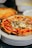 A freshly baked pizza topped with green olives, red bell pepper strips, pieces of sausage or meat, and an egg at the center. The pizza has a golden-brown crust and is placed on a stack of white plates. A blurred background suggests a kitchen or dining setting.