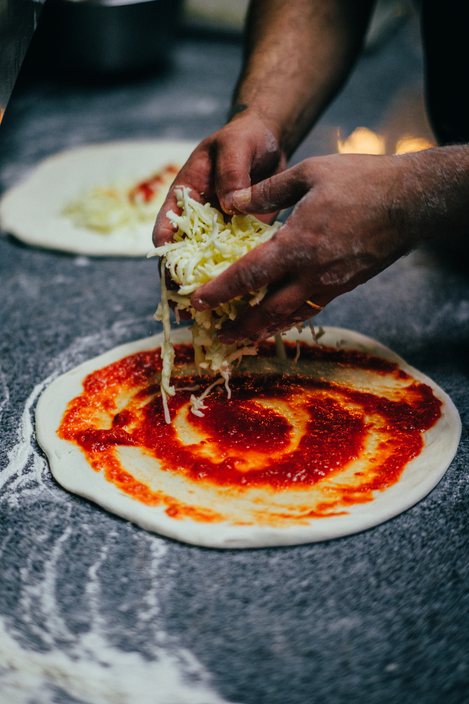 Hands laying a fistful of shredded mozzarella onto a freshly sauced pizza base