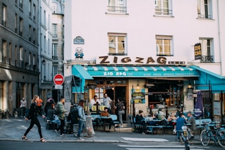 A bustling street cafe with people sitting at outdoor tables. The building has a large sign that reads 'Zigzag Café' and features an 8-bit style artwork on the corner of the wall. The surrounding is urban with narrow streets and people walking by.