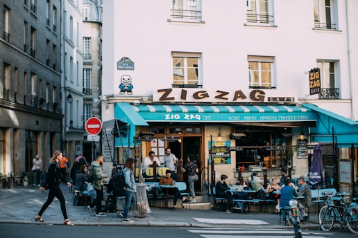 A bustling street cafe with people sitting at outdoor tables. The building has a large sign that reads 'Zigzag Café' and features an 8-bit style artwork on the corner of the wall. The surrounding is urban with narrow streets and people walking by.