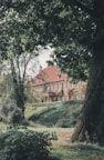 A large, rustic brick building with a red roof is partly obscured by lush green trees and foliage. The setting appears tranquil and secluded, with a well-maintained garden and a sloping lawn leading up to the structure.