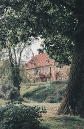 A large, rustic brick building with a red roof is partly obscured by lush green trees and foliage. The setting appears tranquil and secluded, with a well-maintained garden and a sloping lawn leading up to the structure.
