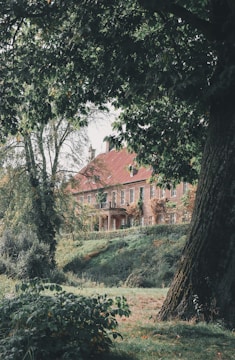 A large, rustic brick building with a red roof is partly obscured by lush green trees and foliage. The setting appears tranquil and secluded, with a well-maintained garden and a sloping lawn leading up to the structure.