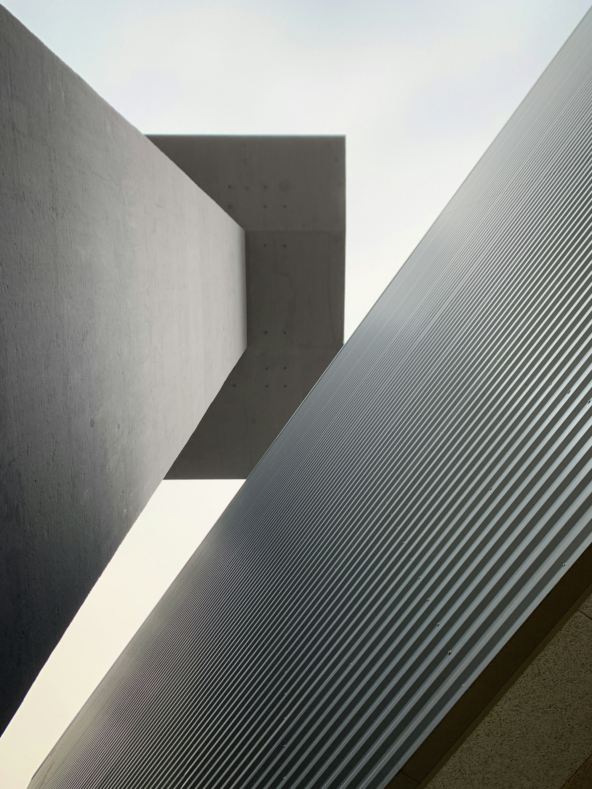 A geometric composition featuring large architectural structures with a focus on sharp lines and contrasting surfaces. The image captures a minimalist aesthetic with one structure having a smooth, flat surface and the other featuring a ribbed texture.