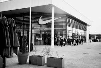 A black-and-white image of a modern building with large windows displaying the Nike logo prominently. There is a group of people standing outside the entrance of the store, with some holding shopping bags and a stroller in the crowd. In the foreground, there are large potted plants and several closed umbrellas.