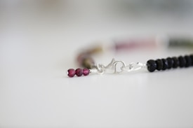 A delicate bracelet featuring small, glossy beads in a gradient of dark to light colors. The beads are linked together by a silver clasp, which lies against a softly focused, neutral background.