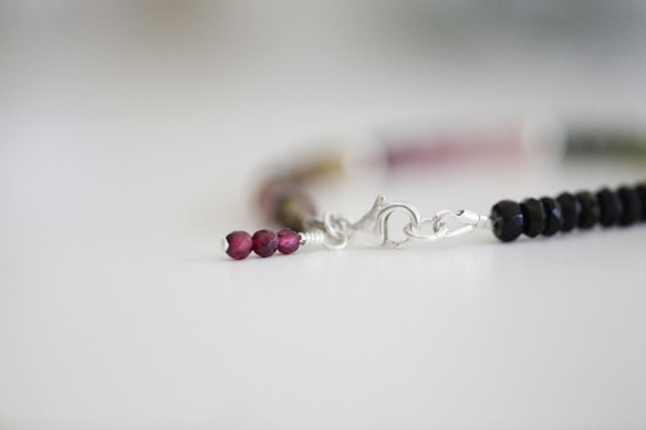 A delicate bracelet featuring small, glossy beads in a gradient of dark to light colors. The beads are linked together by a silver clasp, which lies against a softly focused, neutral background.