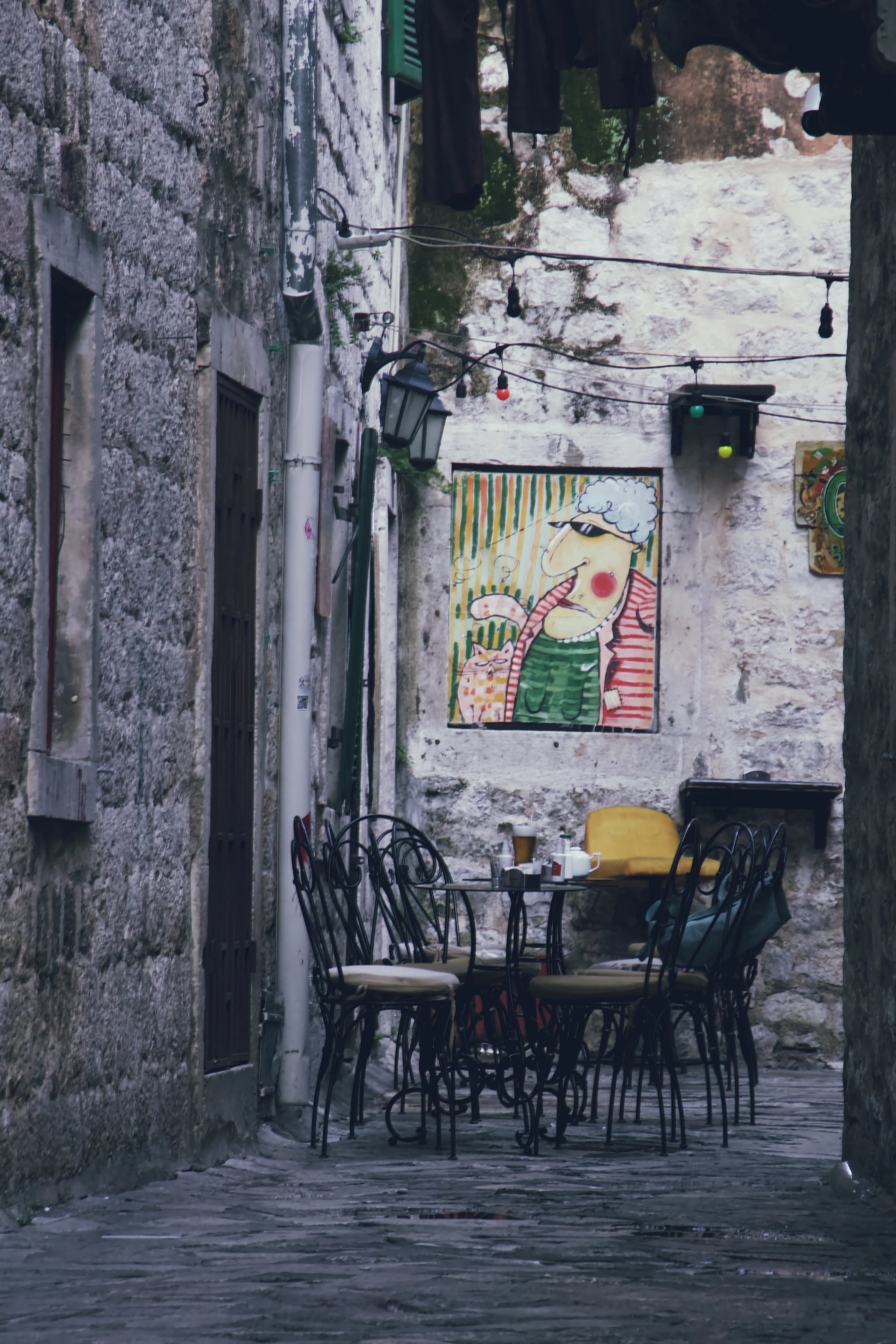 A cafe table set out in a narrow stone alley in Kotor's old town, a tall glass and a teapot on the marble top, wrought-iron chairs pulled around it, a naive painting hung on the wall and a string of bulbs overhead.