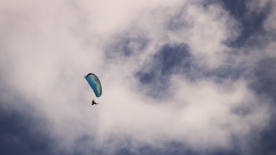 A person is paragliding through a partly cloudy sky, creating a sense of freedom and adventure. The parachute is blue-green and stands out against the backdrop of white and gray clouds.