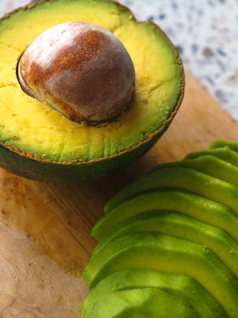 A halved avocado with the pit exposed, resting on a wooden surface. Next to it are several slices of avocado arranged neatly. The texture of the avocado flesh is smooth and creamy, with a vibrant green color.