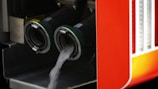 Two large industrial hoses are attached to a structure, with clear liquid pouring out of one. The bright orange surface alongside the hoses suggests a vehicle, possibly related to fire-fighting or industrial use.