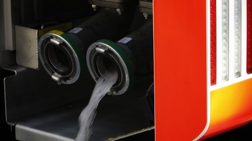 Two large industrial hoses are attached to a structure, with clear liquid pouring out of one. The bright orange surface alongside the hoses suggests a vehicle, possibly related to fire-fighting or industrial use.