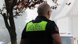 A person with a shaved head wearing a dark police uniform with the word 'POLIZEI' in large, bold letters on the back. The person is seen from behind, and there is a tree nearby with some foliage. In the background, there is a building and part of a police vehicle that has a blue light on top.