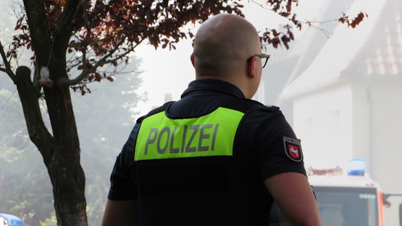 A person with a shaved head wearing a dark police uniform with the word 'POLIZEI' in large, bold letters on the back. The person is seen from behind, and there is a tree nearby with some foliage. In the background, there is a building and part of a police vehicle that has a blue light on top.
