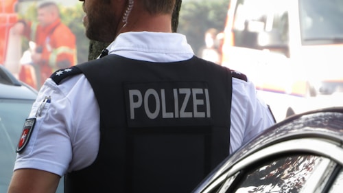 A police officer, identifiable by the label 'POLIZEI' on the back of the vest, stands beside a car. The officer is wearing a white shirt under a black vest with a patch depicting a badge on the sleeve. In the background, there are blurred figures, possibly emergency personnel, wearing orange uniforms, as well as a vehicle that resembles an ambulance.