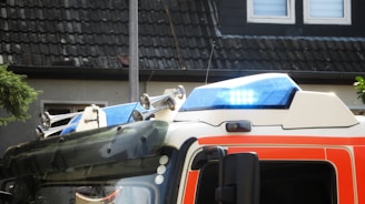 The image captures the top part of an emergency vehicle with a focus on its blue emergency lights and sirens. The vehicle features a white and orange color scheme. Behind it, there is a dark-tiled roof and a wall with windows that suggest a residential area.