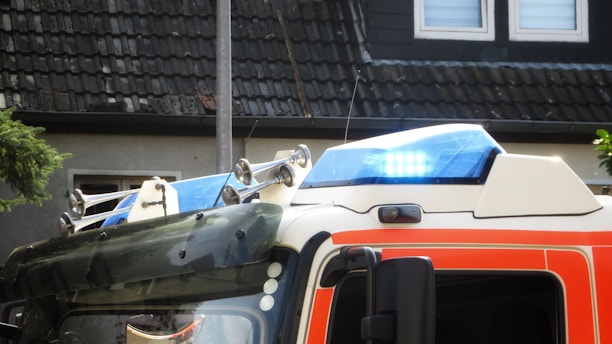 The image captures the top part of an emergency vehicle with a focus on its blue emergency lights and sirens. The vehicle features a white and orange color scheme. Behind it, there is a dark-tiled roof and a wall with windows that suggest a residential area.